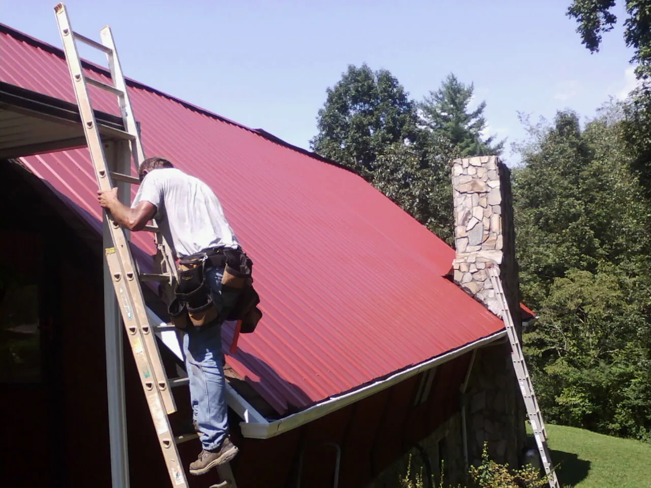 Roofer on a ladder beside a red metal roof and stone chimney — Soffit & Fascia Repair by Roofing Brothers in Woodbine